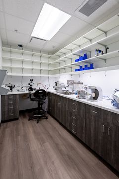 This image of the dental lab workroom at Chroma Dental Studio shows dark lower cabinetry, white counters, open shelving, lab equipment, and bright ceiling lights.