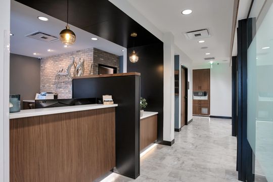 A polished view of the Arvada Dental Center reception area and hallway featuring a wood and black front desk, pendant lighting, brick accents, glass treatment room doors, and modern interior finishes.