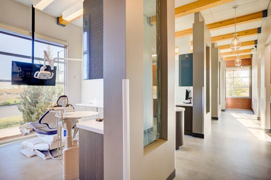 This image of the treatment hallway with dental chair at Meridian Periodontics of Central Park shows a dental chair, wall monitor, glass partitions, wood ceiling beams, and corridor views.