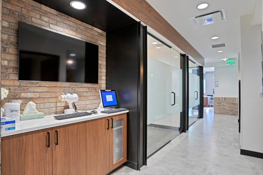 A modern clinical hallway at Arvada Dental Center in Colorado featuring brick accents, wood cabinetry, glass treatment room doors, digital equipment, and a clean contemporary dental office layout.