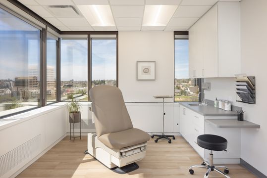 This image of the exam room with city view at Core Dermatology shows an exam chair, large windows, city view, procedure lighting, wall art, and clean counter space.