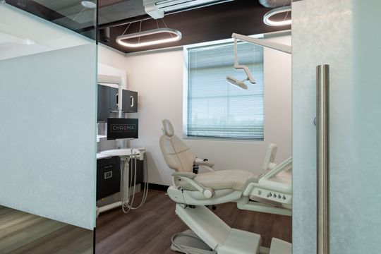 This image of the glass-walled dental operatory at Chroma Dental Studio shows a dental chair behind frosted glass, wall monitor, window shade, and clean treatment room finishes.