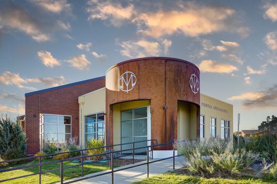 This image of the exterior at sunset at Meridian Periodontics of Central Park shows the dental specialists building, warm evening sky, curved entry volume, windows, and landscape planting.