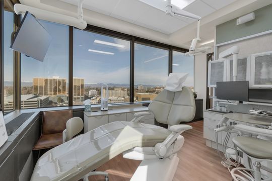 A bright treatment room at Bridge Creek Dentistry with a patient chair, dental equipment, large corner windows, city and mountain views, and modern clinical cabinetry.
