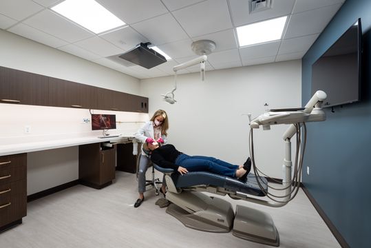 This image of the dental treatment room with patient at Collective Health shows a dental chair, patient, overhead dental equipment, wall monitor, and blue accent wall.