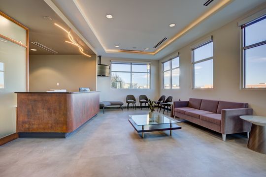 This image of the waiting area and front desk at Meridian Periodontics of Central Park shows a purple sofa, coffee table, reception desk, pendant lighting, and bright window wall.