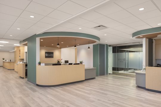 This image of the wide reception lobby at Core Dermatology shows a curved reception desk, green wall panels, pendant lighting, wood ceiling detail, and open lobby flooring.