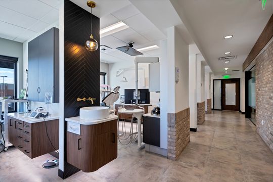 A modern view inside Arvada Dental Center showing a dental operatory, hallway, brick accents, black herringbone tile, pendant lighting, wood cabinetry, and contemporary treatment equipment.