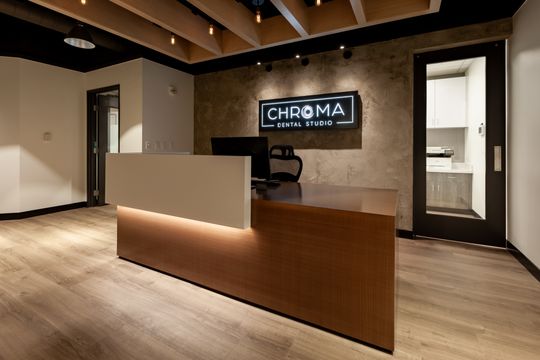 This image of the front desk and entry view at Chroma Dental Studio shows a wood reception counter, branded wall sign, interior doorway, wood flooring, and modern lighting.