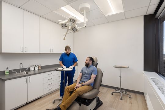 This image of the treatment room consultation at Core Dermatology shows a clinician standing beside a seated patient, procedure light, cabinetry, and clean exam room finishes.