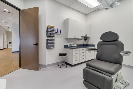 A bright treatment room at Elevation Dermatology featuring an exam chair, clinical cabinetry, task seating, and overhead lighting.