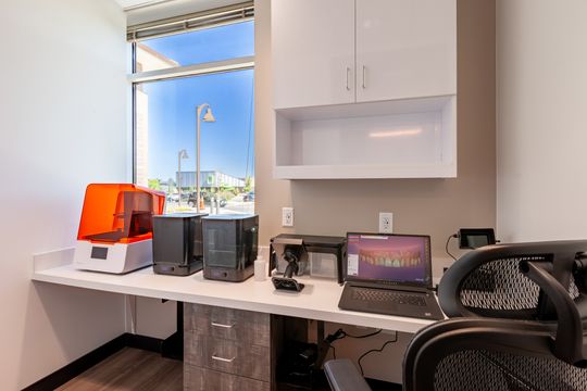This image of the private office desk with window at Chroma Dental Studio shows a workstation, laptop, desk chair, white cabinetry, window view, and desktop equipment.
