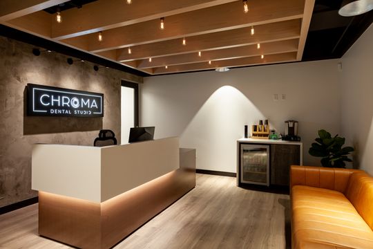 This image of the reception area wide view at Chroma Dental Studio shows a reception counter, branded logo wall, orange lounge seating, beverage station, and warm wood ceiling detail.