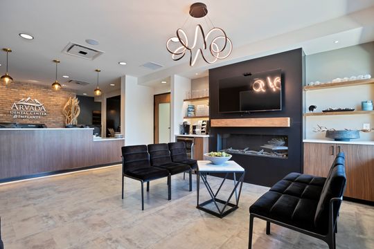 A modern reception and waiting area at Arvada Dental Center in Colorado featuring a wood front desk, brick accent wall, black lounge seating, fireplace feature wall, open shelving, and contemporary lighting.