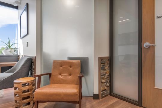 A detailed view of the Bridge Creek Dentistry waiting room featuring a leather lounge chair, side table, fireplace detail, glass office door, and warm interior finishes.