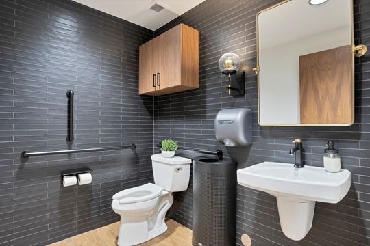 A stylish restroom at Arvada Dental Center featuring dark subway tile, accessibility grab bars, wood cabinetry, a white pedestal sink, gold-framed mirror, and modern black and brass fixtures.