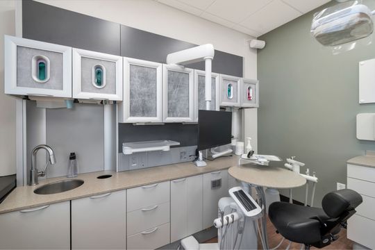 A compact dental operatory at Bridge Creek Dentistry featuring integrated cabinetry, glove storage, clinical sink, patient monitor, treatment chair, and overhead equipment.