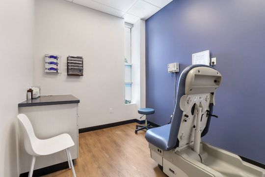 A private exam room at Elevation Dermatology with exam chair, consultation seating, storage, and a calming blue accent wall.