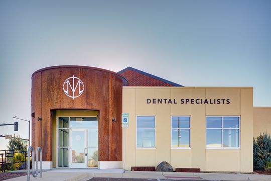This image of the exterior entrance detail at Meridian Periodontics of Central Park shows front signage, curved wood entry, cream wall panels, windows, and accessible parking area.