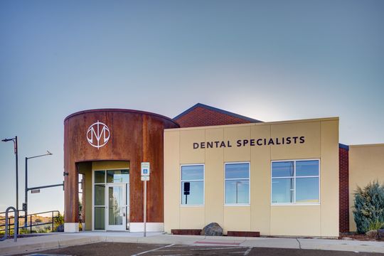 This image of the front entry exterior at Meridian Periodontics of Central Park shows a curved wood entrance, glass door, windows, landscaping, and clear sky.