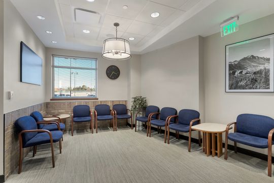 This image of the waiting room seating area at Soult and Swanson Family Dentistry shows blue waiting chairs, side tables, large window, wall art, and soft neutral finishes.