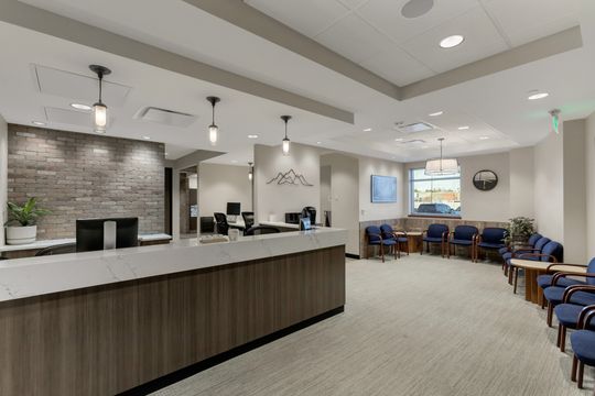 This image of the reception desk and waiting room at Soult and Swanson Family Dentistry shows a long reception counter, stacked stone accent wall, blue waiting chairs, pendant lights, and front office seating.