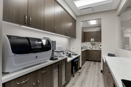 This image of the sterilization lab workspace at Soult and Swanson Family Dentistry shows brown cabinetry, white counters, sterilization equipment, sink, and glass-front storage.